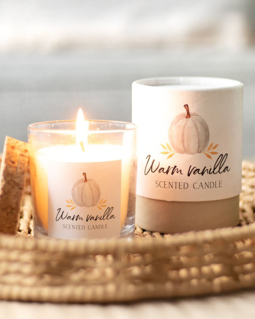 A close-up of the Warm Vanilla scented candle by Rebel Romance, flickering gently in a glass jar. The packaging features a minimalist white pumpkin illustration and warm neutral tones, making it a perfect addition to autumnal home décor.