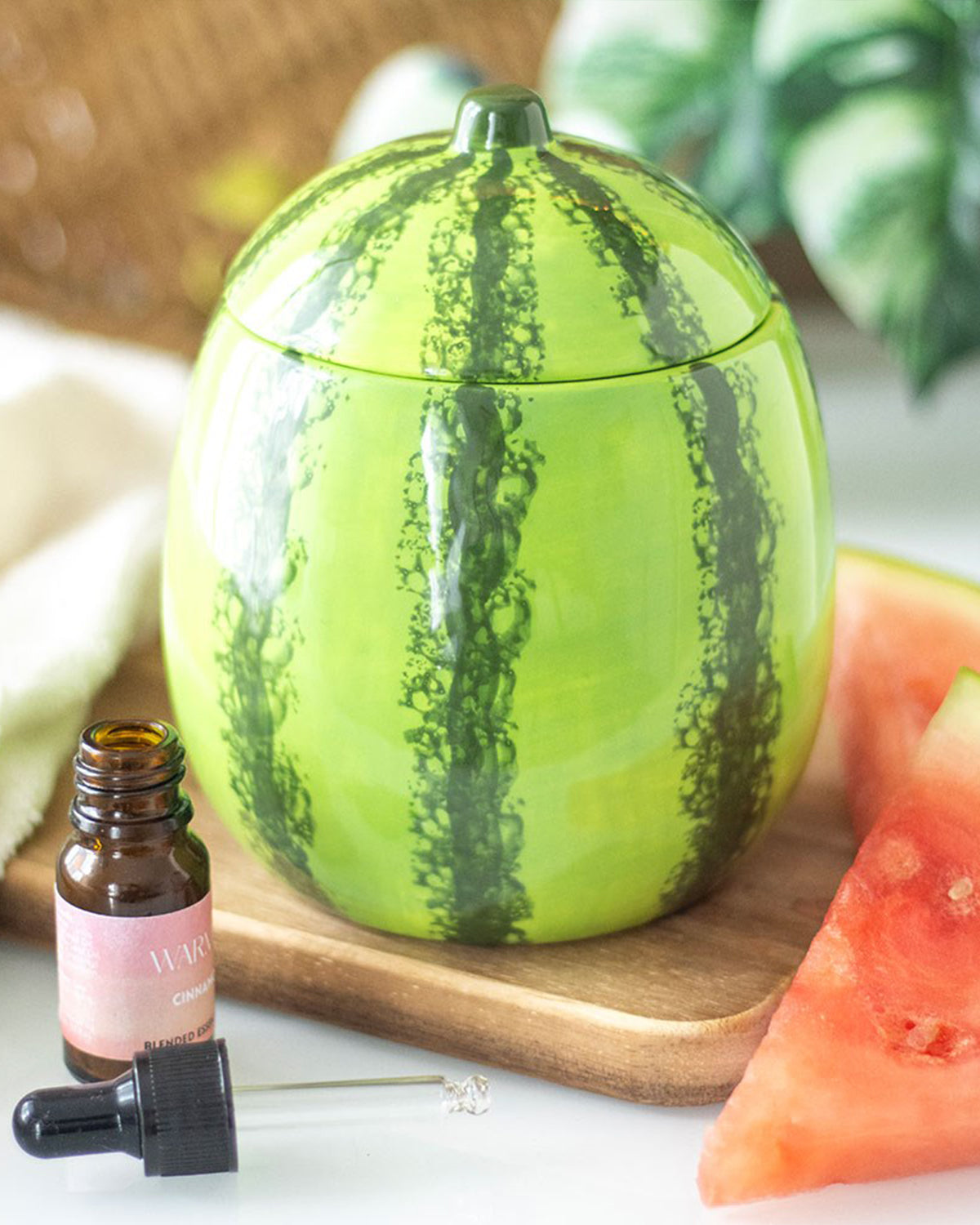 A lifestyle image of the watermelon oil burner styled in a cosy setting with fresh watermelon slices and essential oils. The glossy finish of the ceramic piece reflects light, enhancing its vibrant colours.