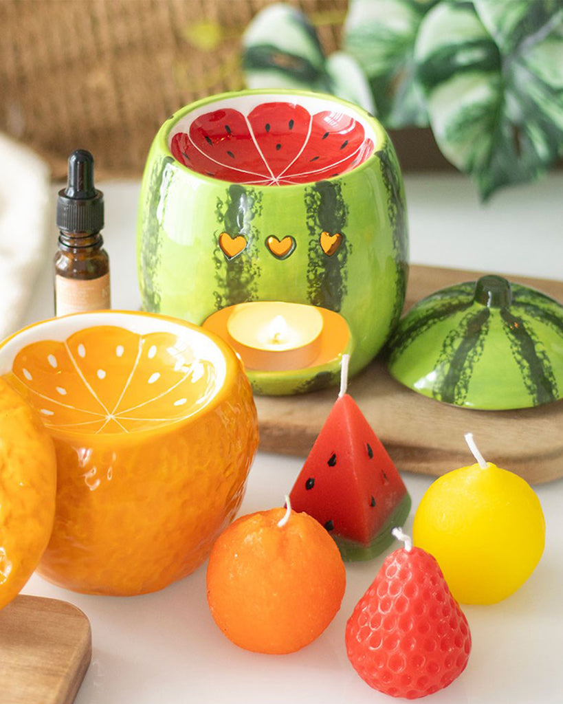 A vibrant ceramic oil burner shaped like a watermelon, featuring a glossy green rind exterior with dark stripes and a bright red interior mimicking the fruit’s flesh. The burner has heart-shaped cutouts for ventilation and a removable lid. It is displayed on a wooden board with essential oils, candles, and fruit slices around it.