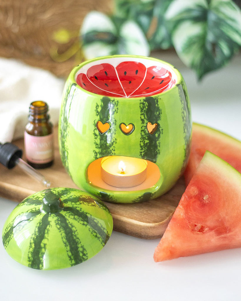 A side view of the oil burner in use, with a lit tealight candle inside, casting a warm glow through the heart-shaped cutouts. The lid is placed next to the burner, and a small dropper bottle of fragrance oil and fresh watermelon slices are included in the setup.