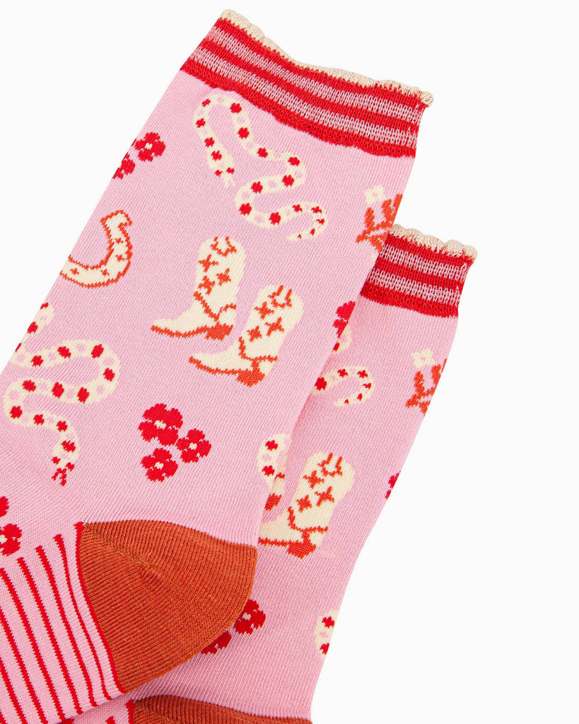 A pair of pink bamboo socks with a wild west theme featuring cowboy boots with heart details, red and cream rattlesnakes, horseshoes, and floral accents. The socks have a striped scalloped cuff, a burnt-orange heel, and red toes.