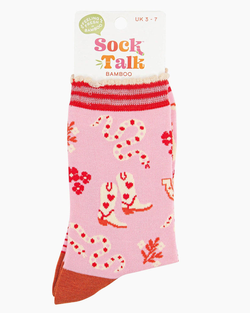 A pair of pink bamboo socks with a wild west theme featuring cowboy boots with heart details, red and cream rattlesnakes, horseshoes, and floral accents. The socks have a striped scalloped cuff, a burnt-orange heel, and red toes.