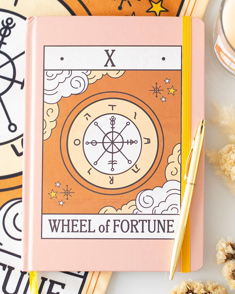 A5 pink vintage tarot notebook featuring the Wheel of Fortune design, with a matching tea towel and candle in the background. Includes yellow elastic strap and gold foil page edges.