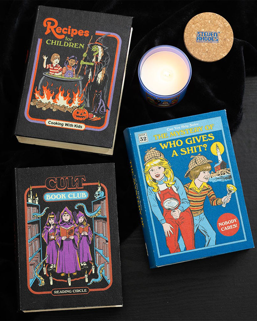 Three book storage boxes with colourful retro inspired covers with dark humour designed by Steven Rhodes on a dark surface, which is lit by a candle.