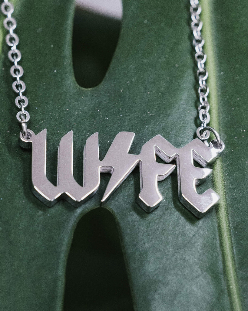 Close-up of a silver metal necklace with the word “WIFE” in a bold, gothic font, featuring a lightning bolt through the middle, displayed on a green monstera leaf background.