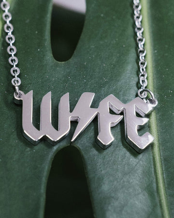 Close-up of a silver metal necklace with the word “WIFE” in a bold, gothic font, featuring a lightning bolt through the middle, displayed on a green monstera leaf background.