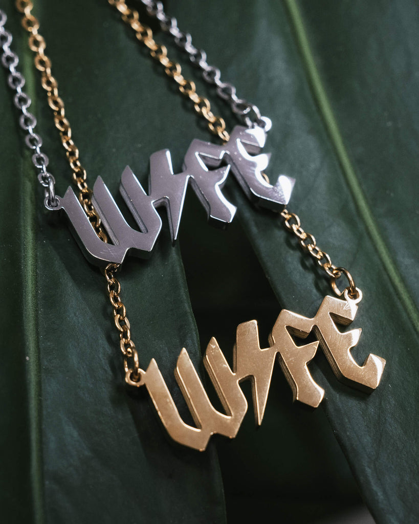 Two “WIFE” necklaces displayed on a green monstera leaf, one in silver and the other in gold, both featuring a bold gothic font and a lightning bolt through the word, with their matching chains attached.