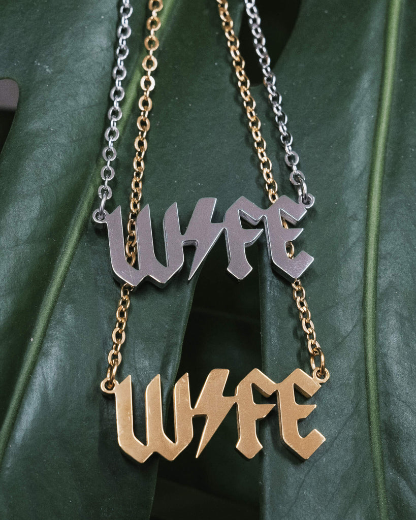 Two “WIFE” necklaces displayed on a green monstera leaf, one in silver and the other in gold, both featuring a bold gothic font and a lightning bolt through the word, with their matching chains attached.