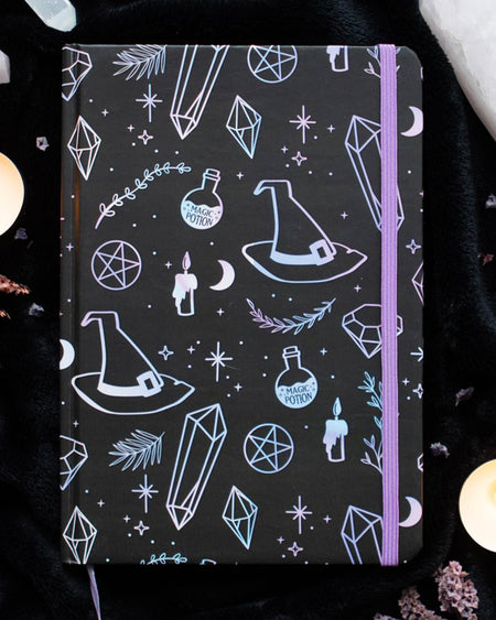 A5 black witchy notebook with holographic pastel illustrations of crystals, potion bottles, moons, and stars. Surrounded by candles and crystals, perfect for spiritual journaling or note-taking.