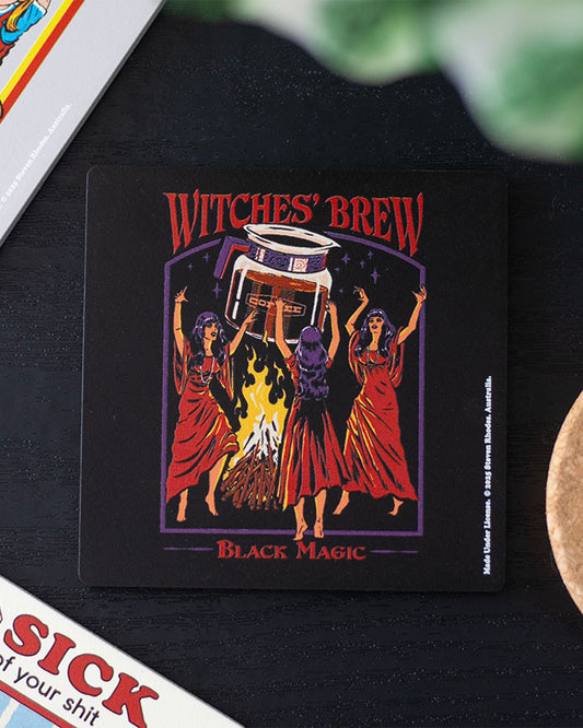 Styled shot of the Rebel Romance Steven Rhodes Witches' Brew Black Magic coaster on a black table. Alternative retro homeware design with three witches worshipping a coffee pot.