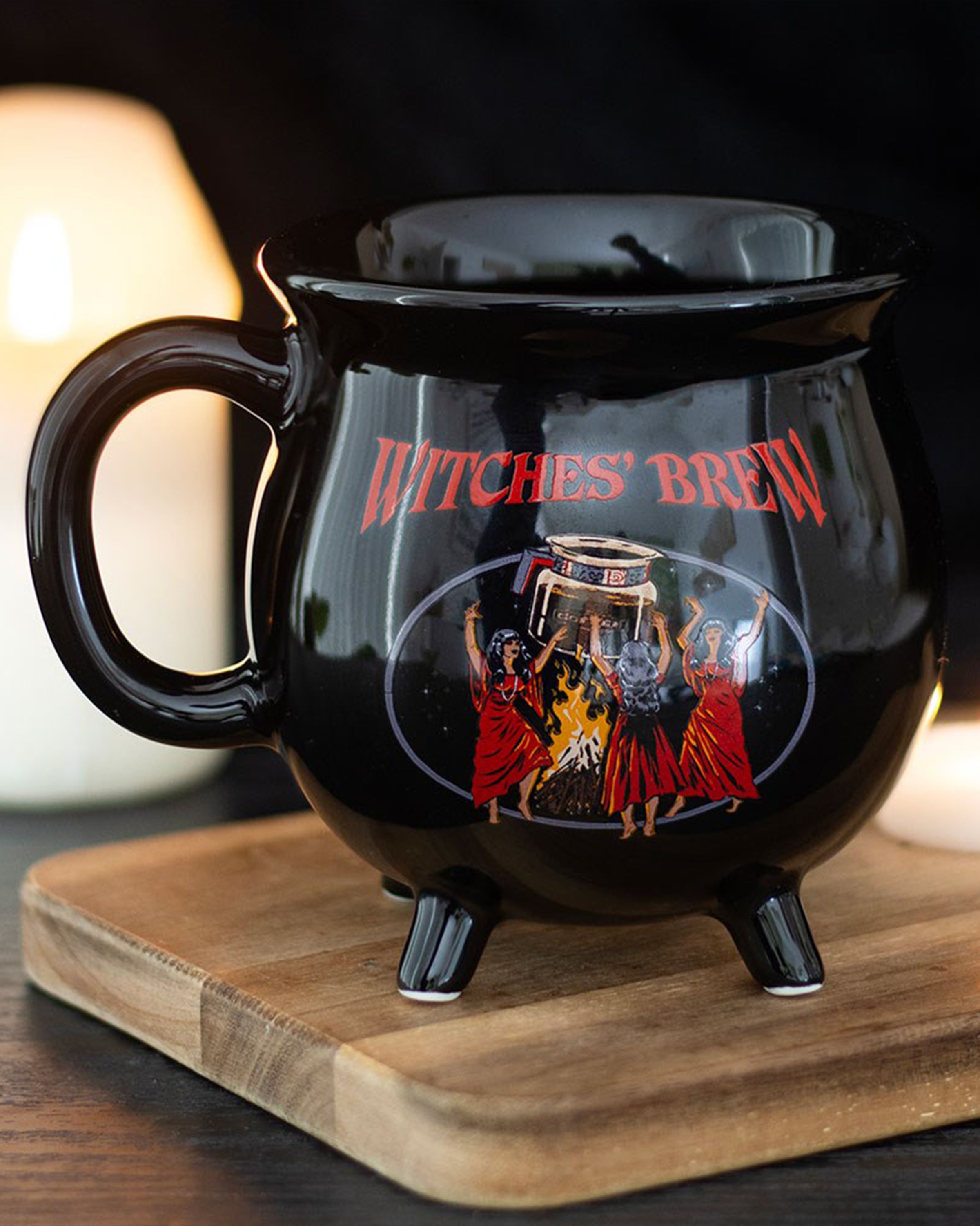 Lifestyle shot of the Rebel Romance Witches' Brew Cauldron Mug by Steven Rhodes on a wooden board with candles in the background. A gothic cauldron-shaped novelty mug, ideal for coffee, tea, or alternative homeware gifts.