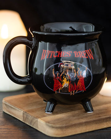 Lifestyle shot of the Rebel Romance Witches' Brew Cauldron Mug by Steven Rhodes on a wooden board with candles in the background. A gothic cauldron-shaped novelty mug, ideal for coffee, tea, or alternative homeware gifts.