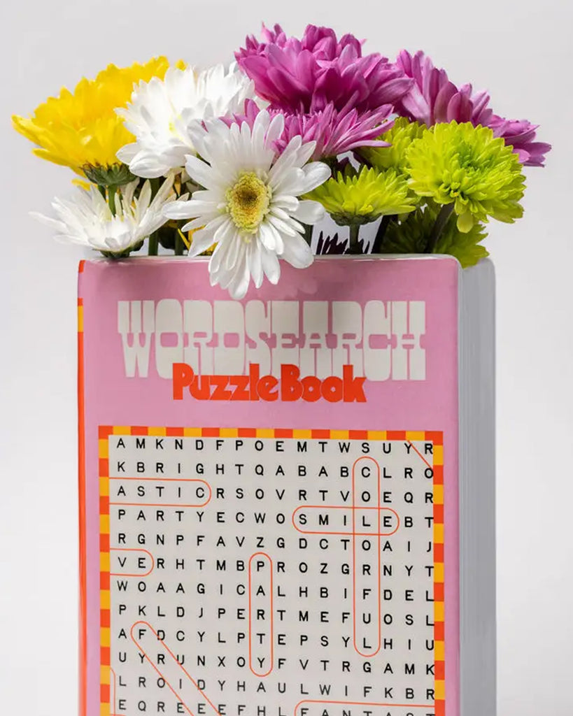 Ceramic vase shaped like a pink word search puzzle book, filled with yellow, white, pink and green flowers, from Rebel Romance, a UK brand celebrating alternative style and unique gifts.