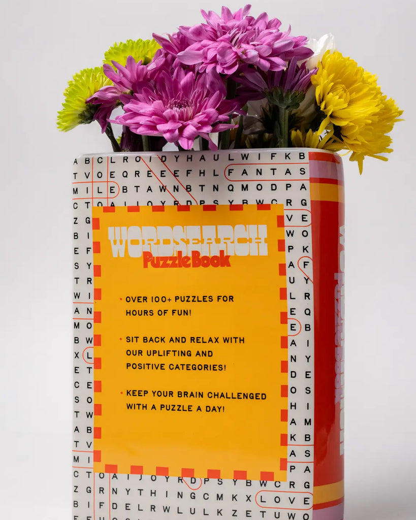 Back view of the Rebel Romance word search vase showing a bright yellow panel with playful text about the benefits of puzzles, surrounded by blooming flowers.