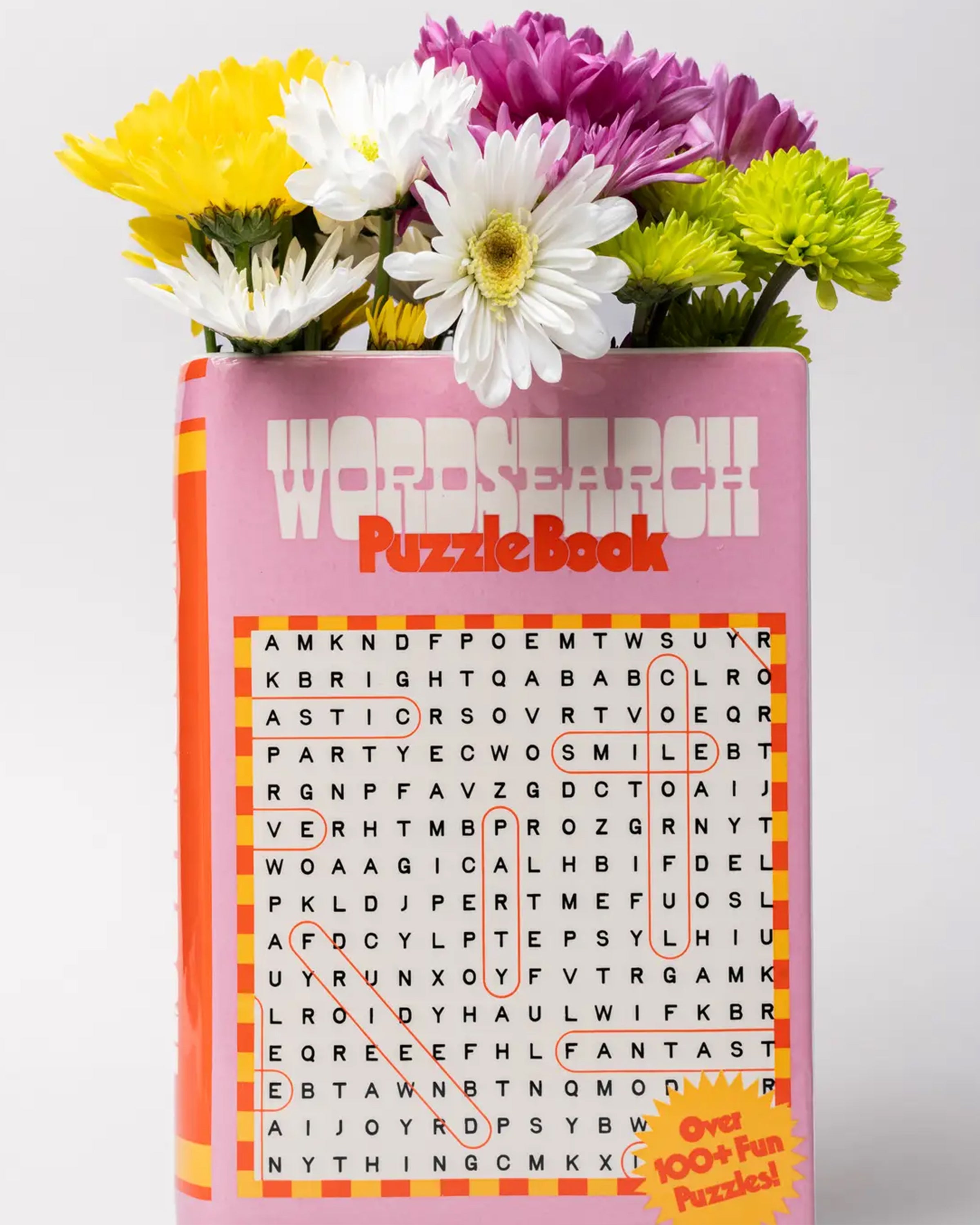 Front view of the Rebel Romance Word Search Puzzle Book Vase styled with colourful fresh flowers, showing a pink retro book cover design and the phrase “Over 100+ Fun Puzzles!”