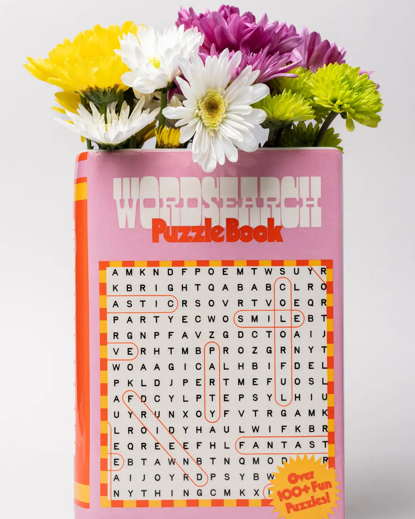 Front view of the Rebel Romance Word Search Puzzle Book Vase styled with colourful fresh flowers, showing a pink retro book cover design and the phrase “Over 100+ Fun Puzzles!”