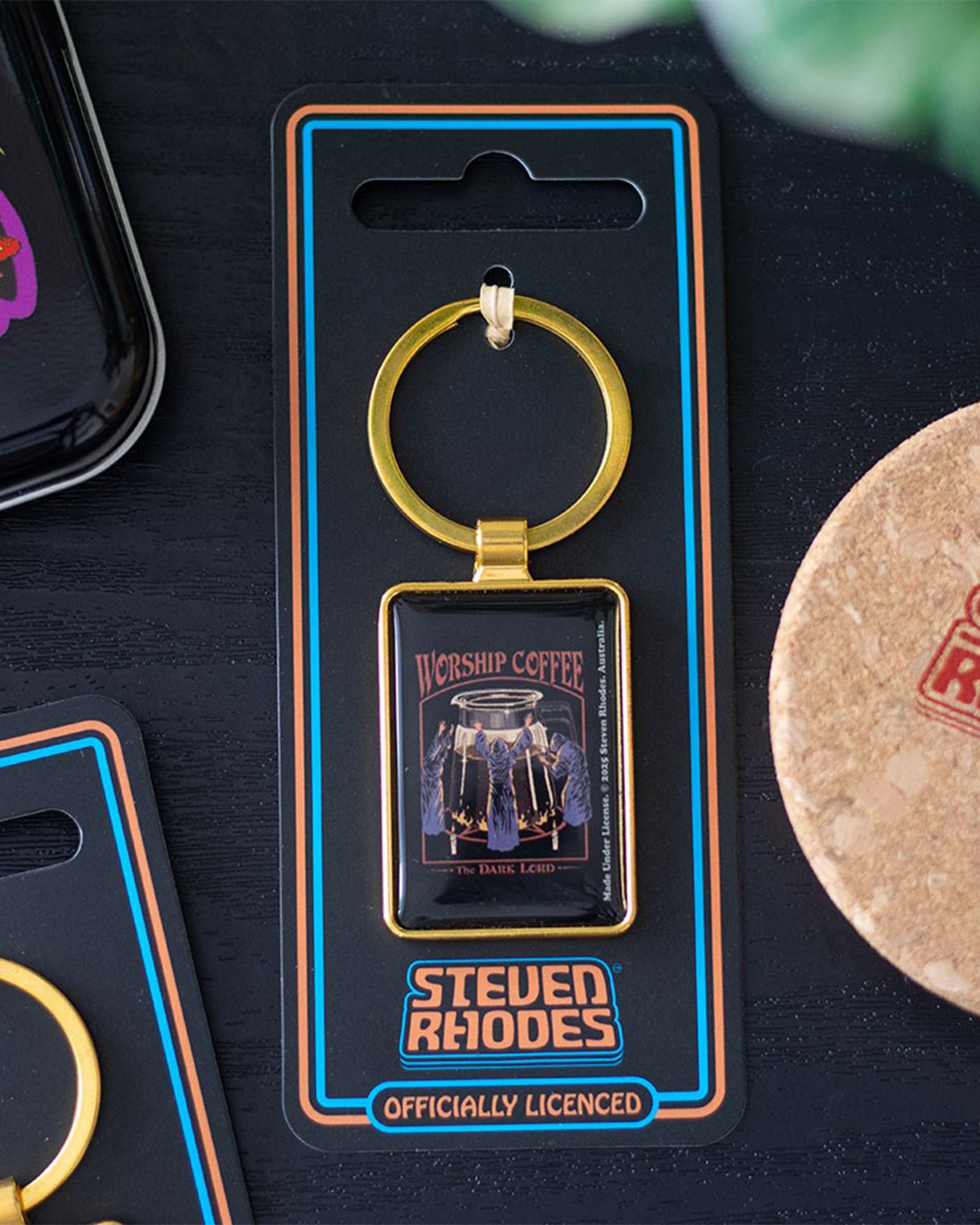 Styled flatlay of the Worship Coffee enamel keyring by Steven Rhodes, on branded packaging beside other retro accessories. Gothic coffee-themed artwork with hooded worshippers around a coffee pot. Sold by Rebel Romance, a UK brand for alternative style and millennial gifts.