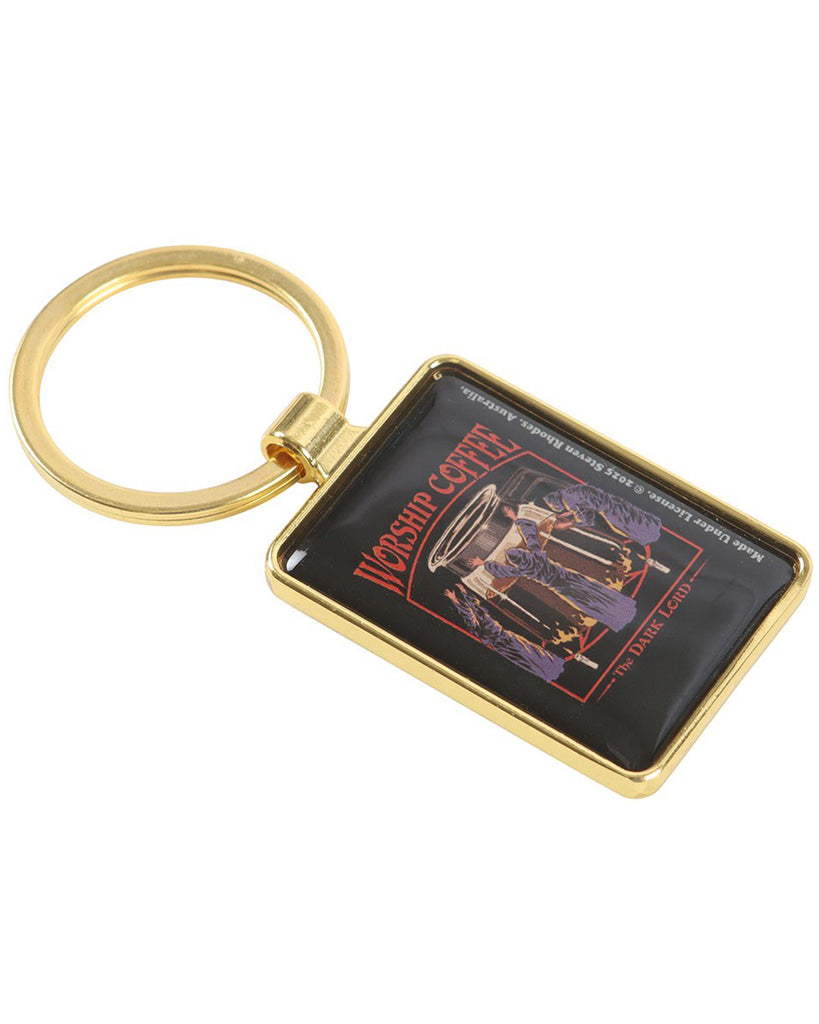 Angled view of the Worship Coffee enamel keyring by Steven Rhodes. The gold keyring frame highlights the dark retro artwork of cult-like figures bowing to a steaming coffee pot. Sold by Rebel Romance, a UK business for alternative fashion, unique gifts, and gothic style accessories.