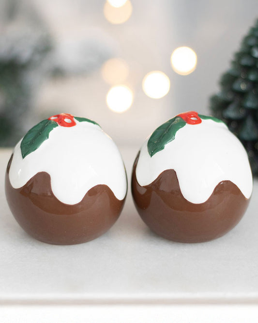 Close-up of two ceramic Christmas pudding salt and pepper shakers with white icing and holly leaf detailing, displayed on a white tray with twinkling lights in the background.