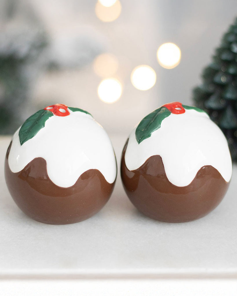 Close-up of two ceramic Christmas pudding salt and pepper shakers with white icing and holly leaf detailing, displayed on a white tray with twinkling lights in the background.