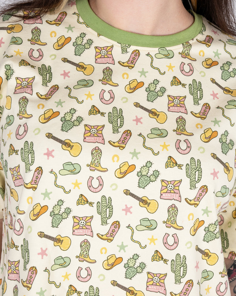 Close-up of the Rebel Romance x Yasmin Cousins Cowboy Cropped Boxy Tee showing its pastel Western print. The cream tee features cowboy boots, hats, guitars, cacti, and smiley ‘Wanted’ signs with green trim on the collar. A fun and ethical fashion choice by Rebel Romance, a UK brand celebrating bold, alternative style for millennials.