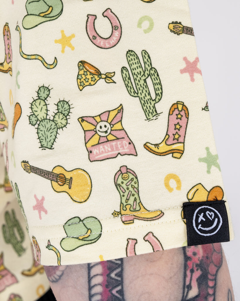 Detailed close-up of the sleeve of the Rebel Romance x Yasmin Cousins Cowboy Cropped Boxy Tee, highlighting the playful cowboy print with boots, hats, cacti, and guitars. The Rebel Romance smiley face tag is visible on the hem, showcasing the small UK brand's quirky and inclusive fashion made for alternative dressers.