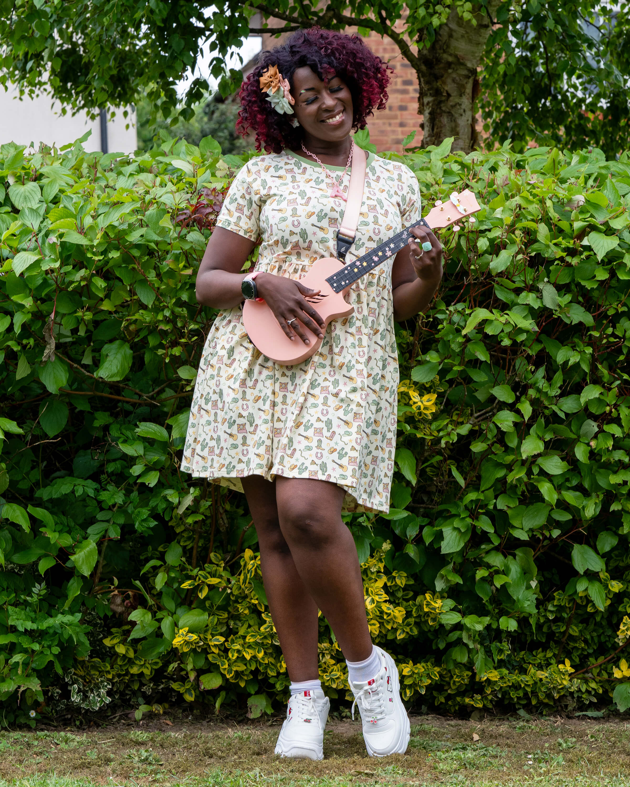 Ayesha plays a pale pink ukulele while wearing the Rebel Romance x Yasmin Cousins Cowboy Stretch Smock Dress. The dress features a fun pastel western print with boots, cacti and guitars. She stands in front of green bushes, styled with chunky trainers and bow hair clips.