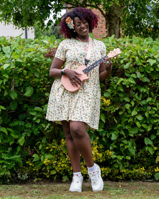Ayesha plays a pale pink ukulele while wearing the Rebel Romance x Yasmin Cousins Cowboy Stretch Smock Dress. The dress features a fun pastel western print with boots, cacti and guitars. She stands in front of green bushes, styled with chunky trainers and bow hair clips.