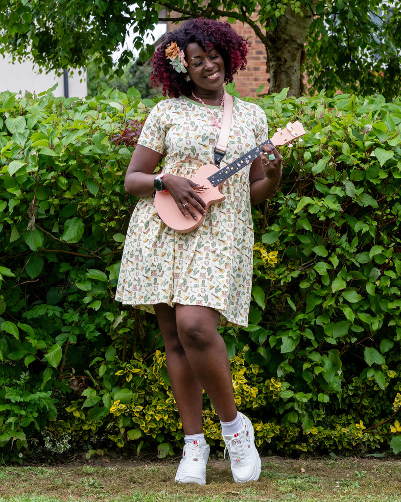 Ayesha plays a pale pink ukulele while wearing the Rebel Romance x Yasmin Cousins Cowboy Stretch Smock Dress. The dress features a fun pastel western print with boots, cacti and guitars. She stands in front of green bushes, styled with chunky trainers and bow hair clips.