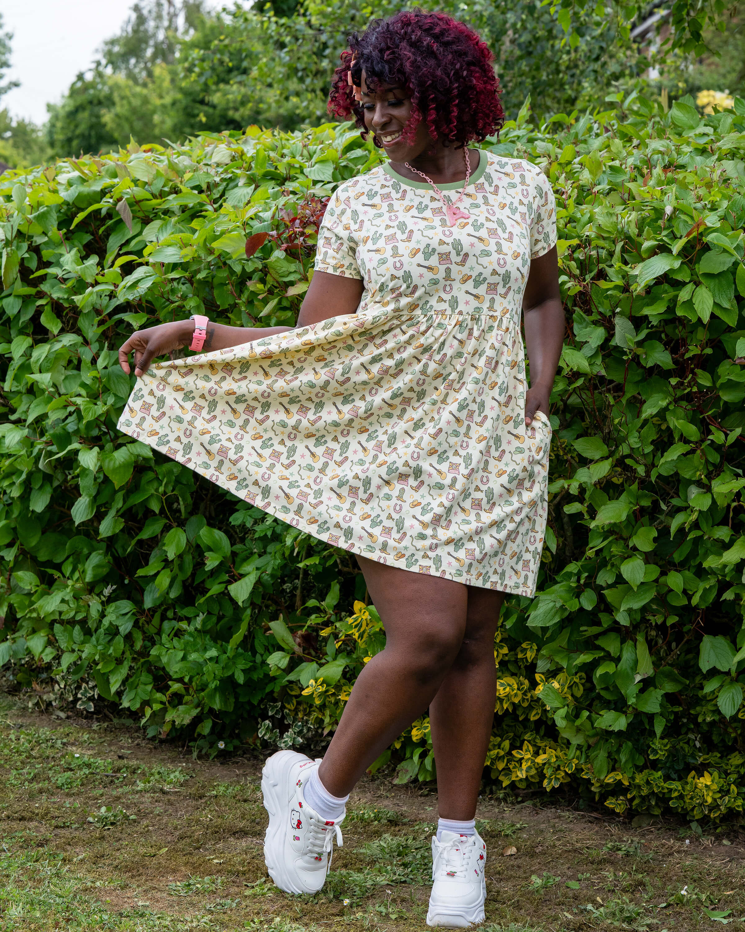 Ayesha poses playfully in the Rebel Romance x Yasmin Cousins Cowboy Stretch Smock Dress, holding the skirt out. The pastel cowgirl print features boots, hats, stars, and cacti. She wears chunky white Hello Kitty trainers and stands in a garden setting.