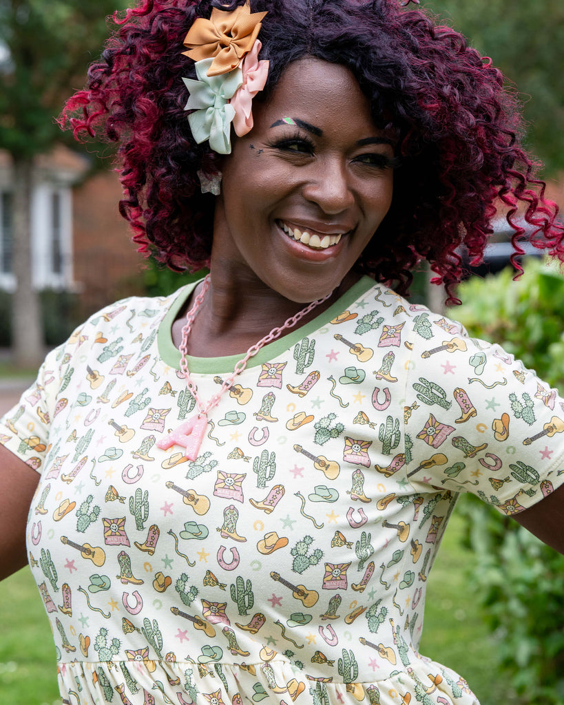 Close-up of Ayesha wearing the Rebel Romance x Yasmin Cousins Cowboy Stretch Smock Dress. The western-themed print includes cowboy boots, guitars, cacti, and stars in muted pastel tones. She smiles brightly with bow hair clips, a pink chain necklace, and colourful makeup.