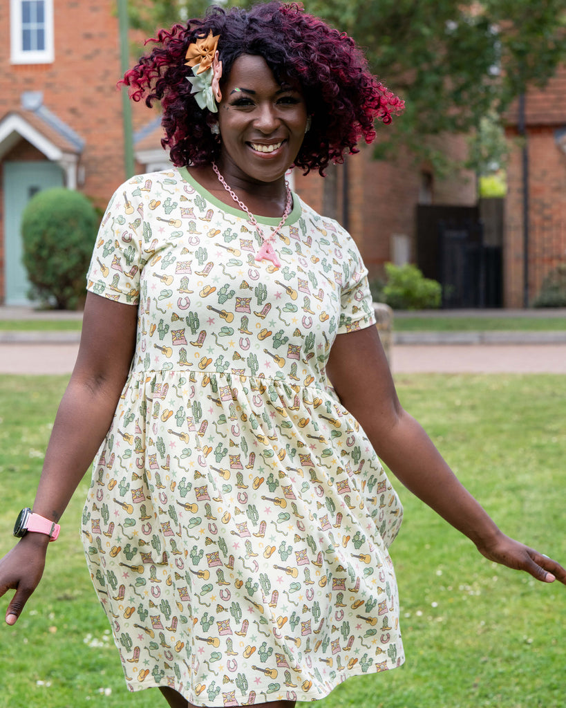 Ayesha wears the Rebel Romance x Yasmin Cousins Cowboy Stretch Smock Dress in a garden setting. The cream dress features a pastel western print with boots, hats, cacti and stars. Ayesha smiles with one hand to her head, showing the playful, comfy silhouette. She wears white Hello Kitty trainers and a pink watch.