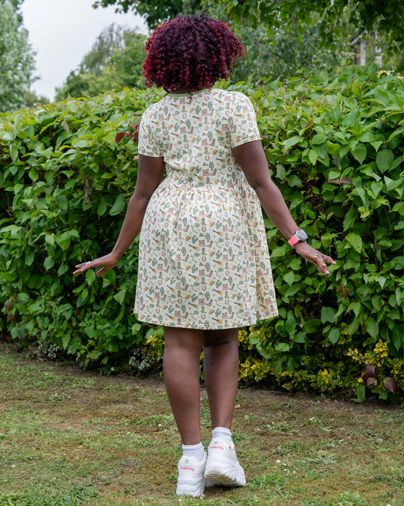 Back view of Ayesha in the Rebel Romance x Yasmin Cousins Cowboy Stretch Smock Dress. The short-sleeved dress gently flares out, showing the pastel cowgirl-themed pattern. She pairs it with white Hello Kitty trainers and stands in front of green bushes outdoors.