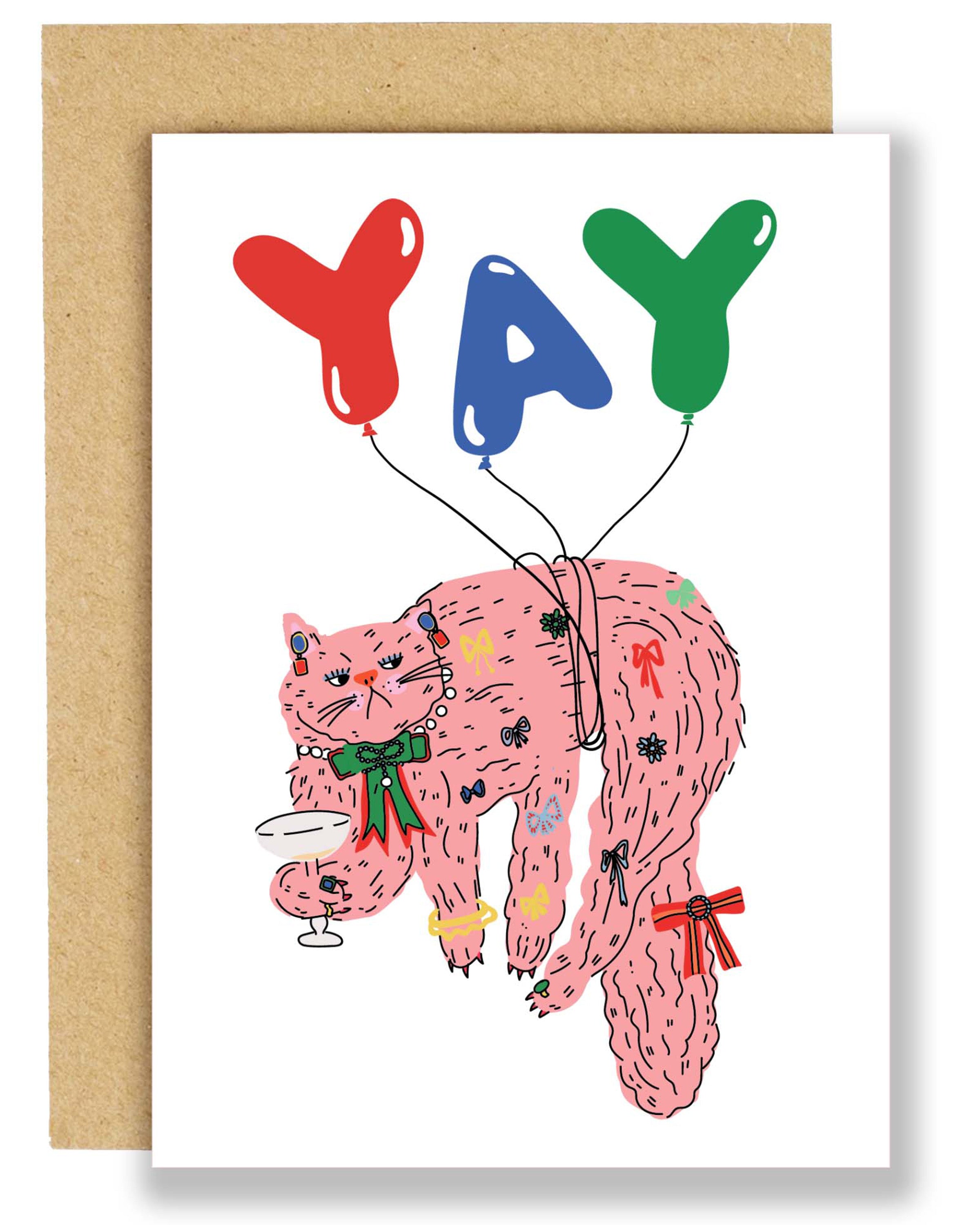 Illustrated birthday card featuring a pink cat covered in bows and holding a cocktail, attached to colourful balloons spelling “YAY,” with a brown Kraft envelope in the background.