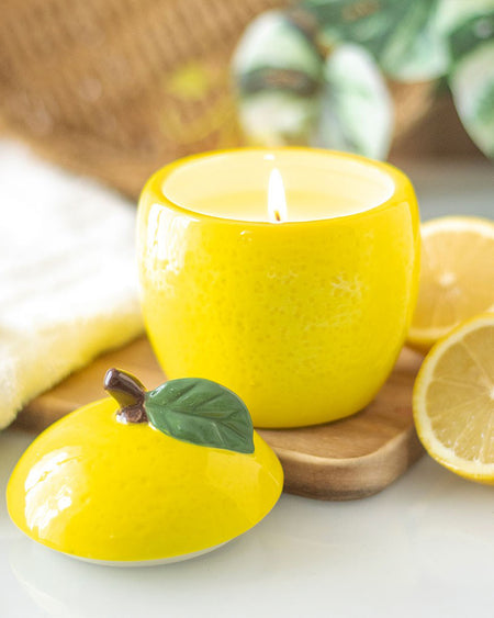 The lemon candle jar lit, emitting a warm glow. The removable ceramic lid rests beside it, showing off its realistic fruit-inspired design.