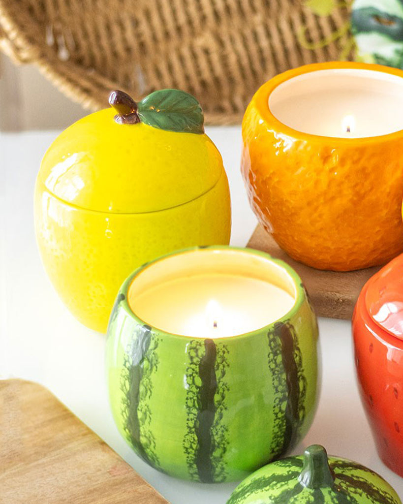 A collection of fruit-shaped candle jars, featuring lemon, orange, and watermelon designs. The ceramic jars have removable lids and textured surfaces, creating a playful and colourful home décor aesthetic.