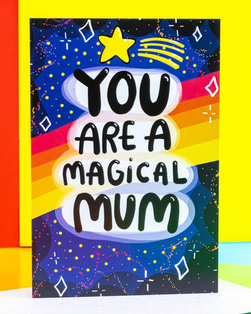 A colourful A6 greeting card by Rebel Romance, a small UK business celebrating alternative style. The card features a bold and playful cosmic design by Katie Abey with the words “You Are A Magical Mum” in chunky black text on a rainbow and starry night background. Ideal for Mother’s Day or just because. Surrounded by bright yellow and red props. Blank inside for your message.