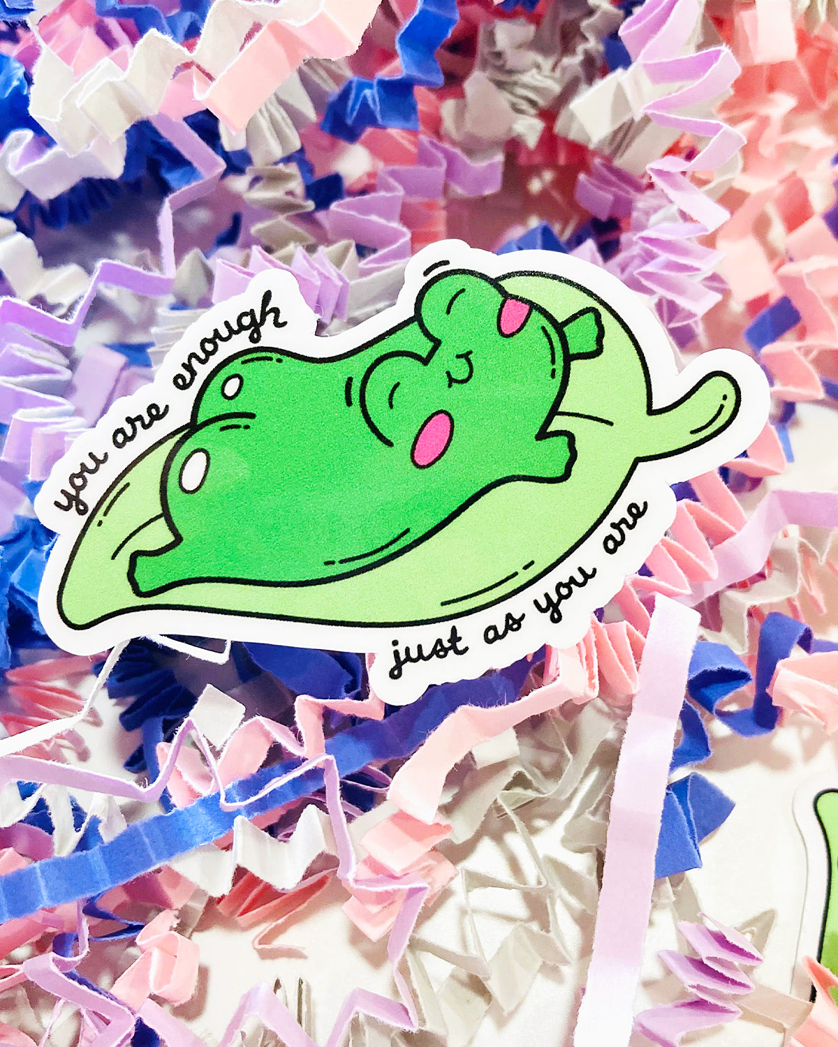 Die cut vinyl sticker from Rebel Romance featuring a happy green frog lying on a lily pad with pink cheeks. Surrounded by pastel shredded paper in pink, purple, and blue tones. The text reads “You are enough just as you are.” A cute, uplifting mental health sticker from a small UK brand celebrating alternative style, self-love, and kindness. Ideal for decorating notebooks, laptops, or gifts.