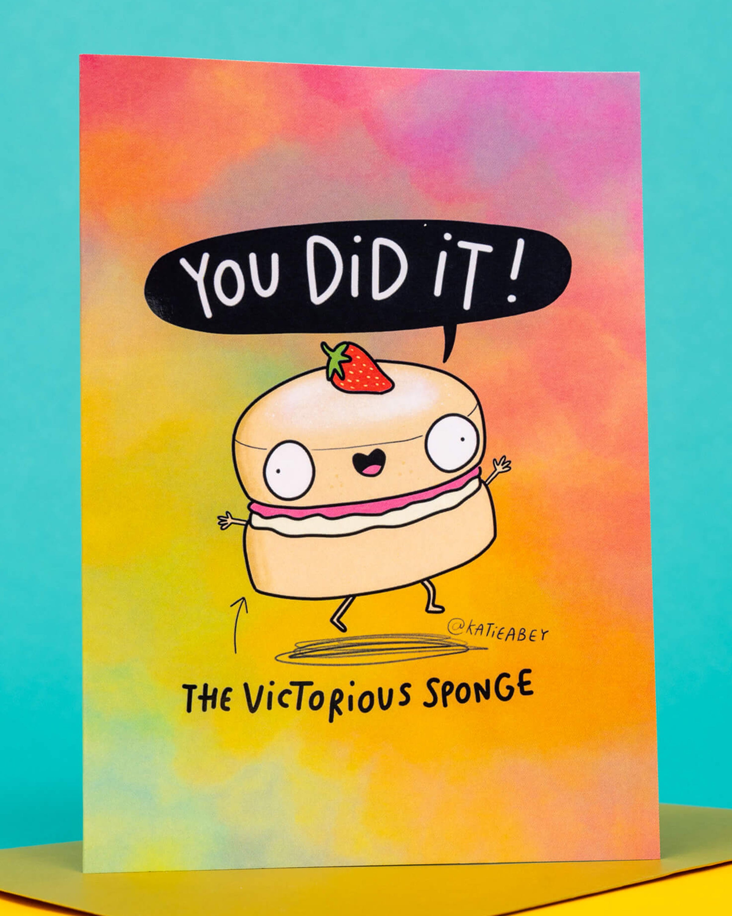 A colourful A6 greeting card from Rebel Romance featuring a cute cartoon sponge cake with big eyes, a strawberry on top, and jam filling, joyfully shouting “You did it!” in a speech bubble. The background is a rainbow gradient of pink, orange and yellow. The text reads “The Victorious Sponge.” Designed by Katie Abey, this funny celebration card is perfect for sending congratulations with quirky charm.