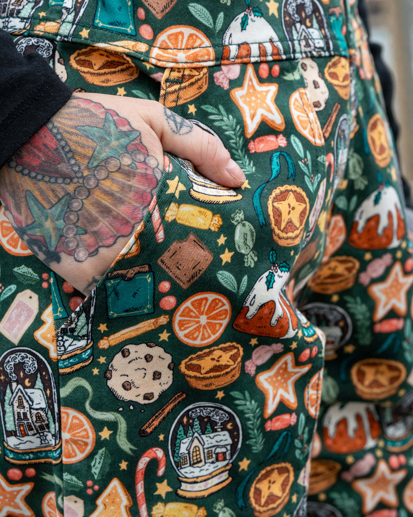 Close-up of the pocket detail on the Rebel Romance Yuletide Treats Dungarees worn by Erin Porter. The fabric shows a bold festive pattern with cookies, oranges, puddings and Christmas trees.