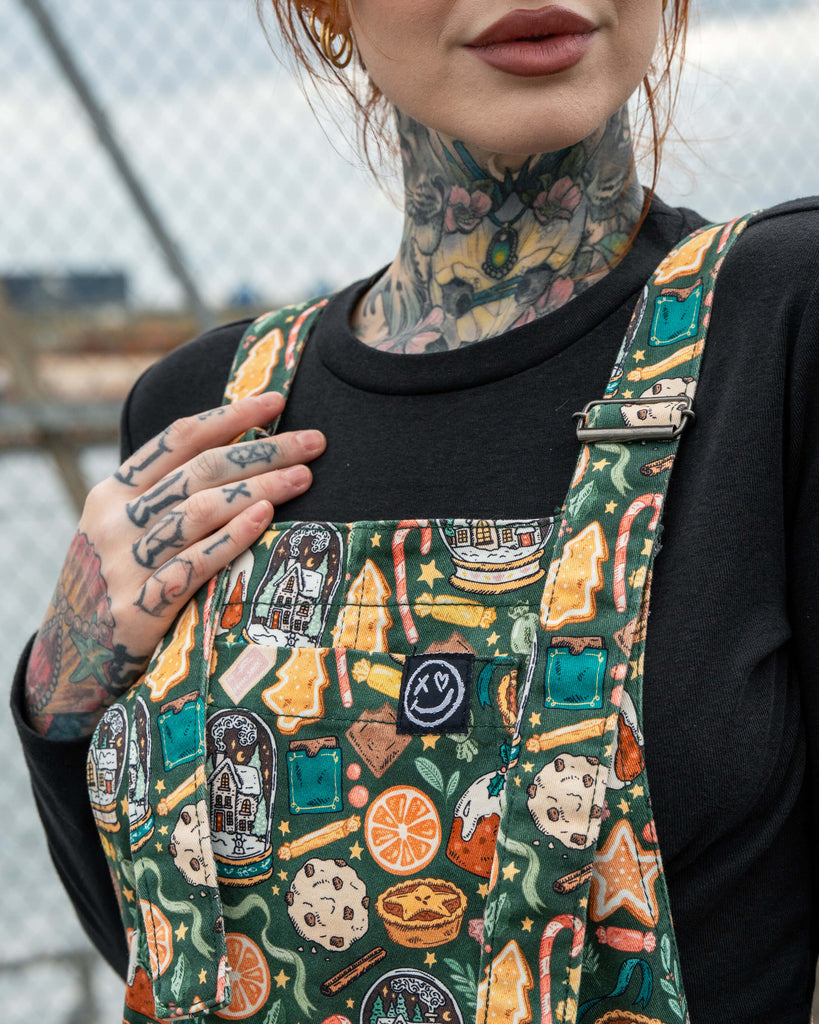 Close-up of the chest detail on the Rebel Romance Yuletide Treats Dungarees worn by Erin Porter. The fabric shows a bold festive pattern with cookies, oranges, puddings and Christmas trees, finished with the Rebel Romance logo on the bib pocket.