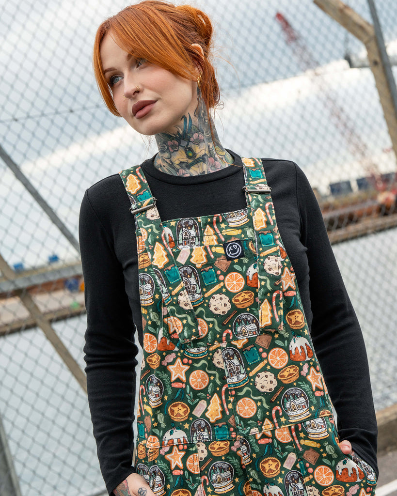 Close-up of Erin Porter wearing the Rebel Romance Yuletide Treats Dungarees. The festive dungarees feature an all-over holiday print with oranges, Christmas puddings, candy canes and gingerbread, styled with a black top for an alternative Christmas look.