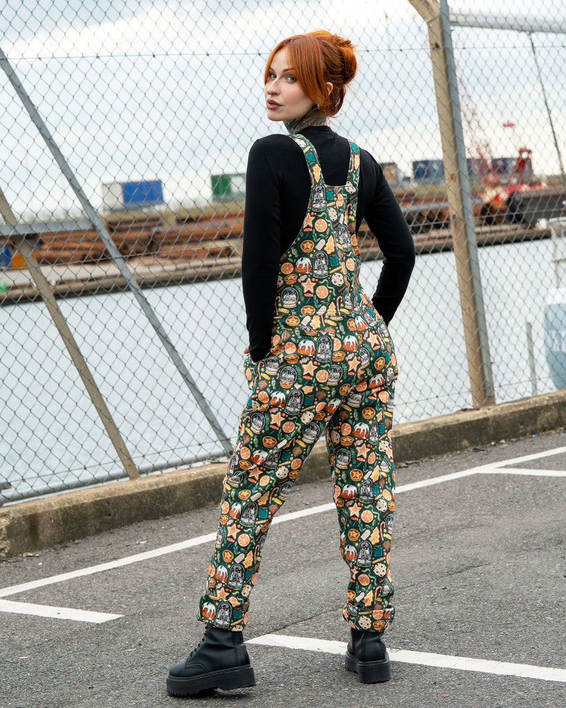 Back view of Erin Porter modelling the Yuletide Treats Dungarees by Rebel Romance. The Christmas-themed dungarees feature a bold print with seasonal treats including gingerbread biscuits, snow globes and mince pies, styled with black boots.
