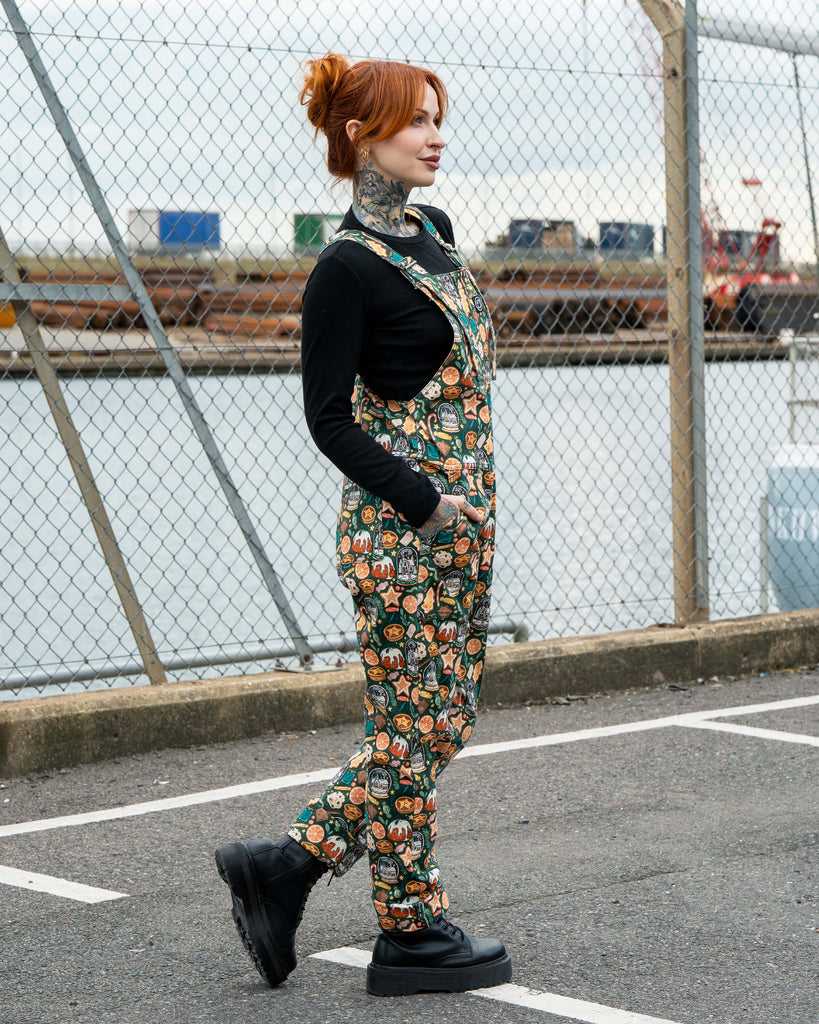 Side profile of Erin Porter in the Rebel Romance Yuletide Treats Dungarees. The alternative festive dungarees feature a green base with gingerbread, puddings and snow globe motifs, styled with a black long-sleeve top and chunky lace-up boots.
