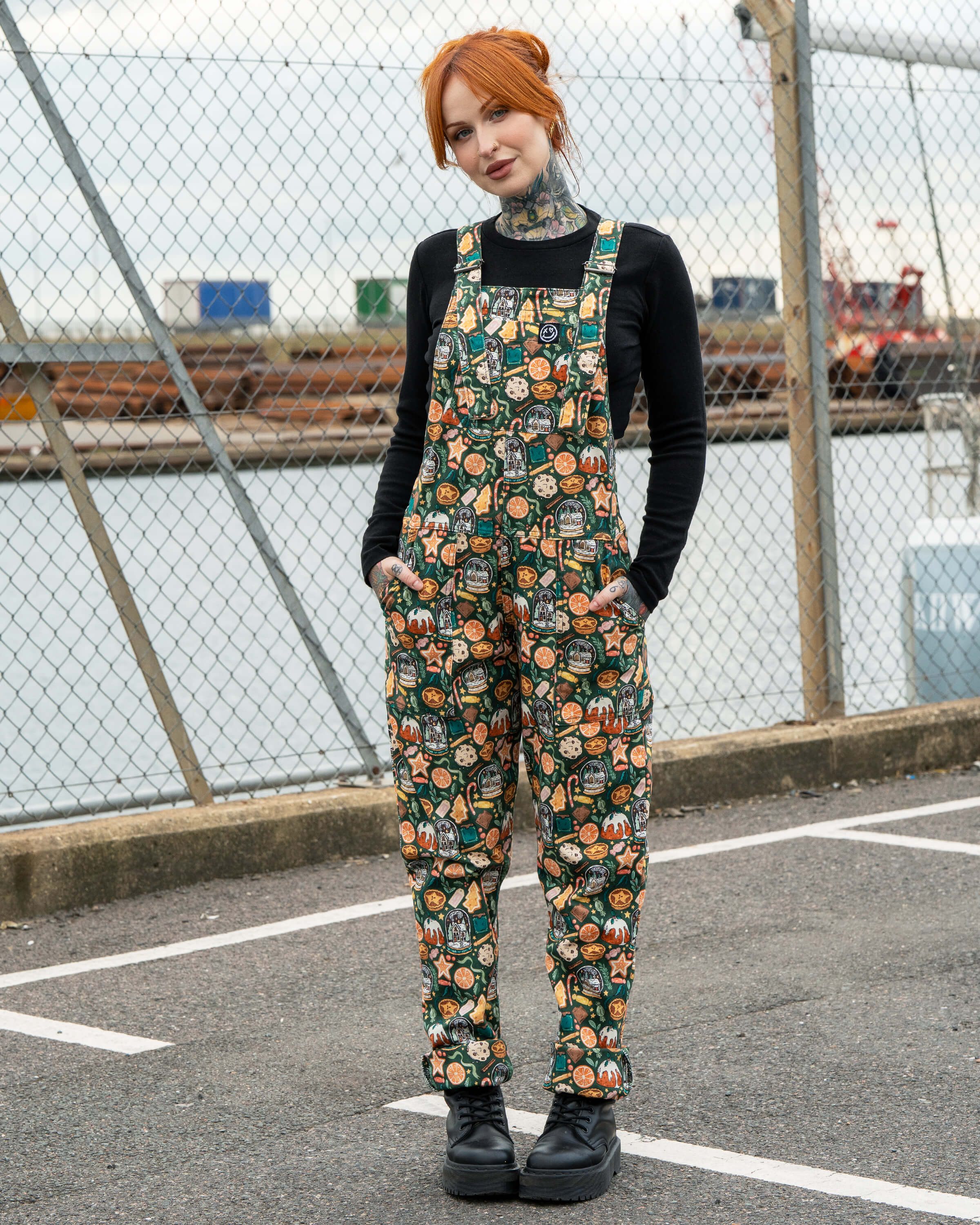 Full-length shot of Erin Porter modelling the Rebel Romance Yuletide Treats Dungarees. The festive all-over print features gingerbread biscuits, snow globes, oranges and puddings, styled with a black top and chunky boots for a playful holiday outfit.
